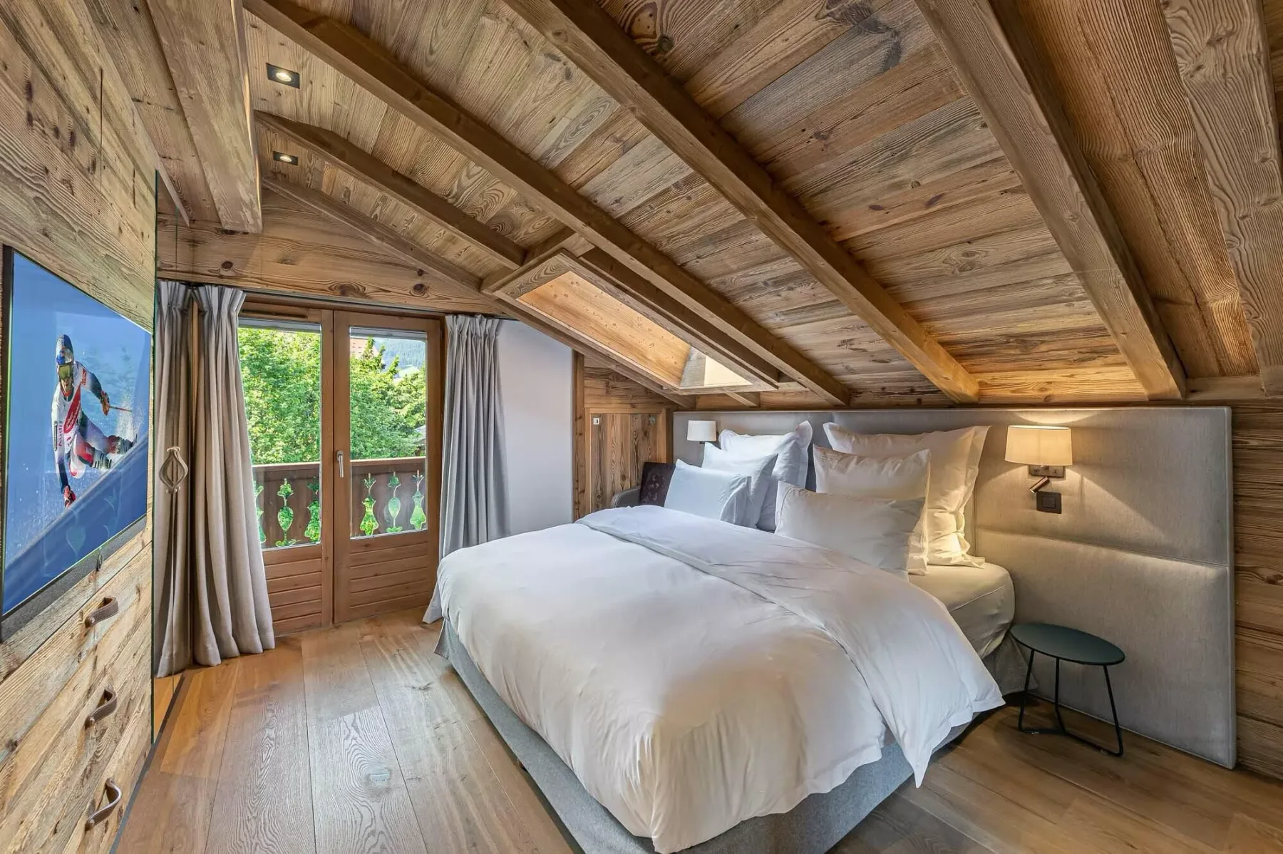 Un dormitorio con una cama grande bajo un techo inclinado de madera y una puerta al balcón.