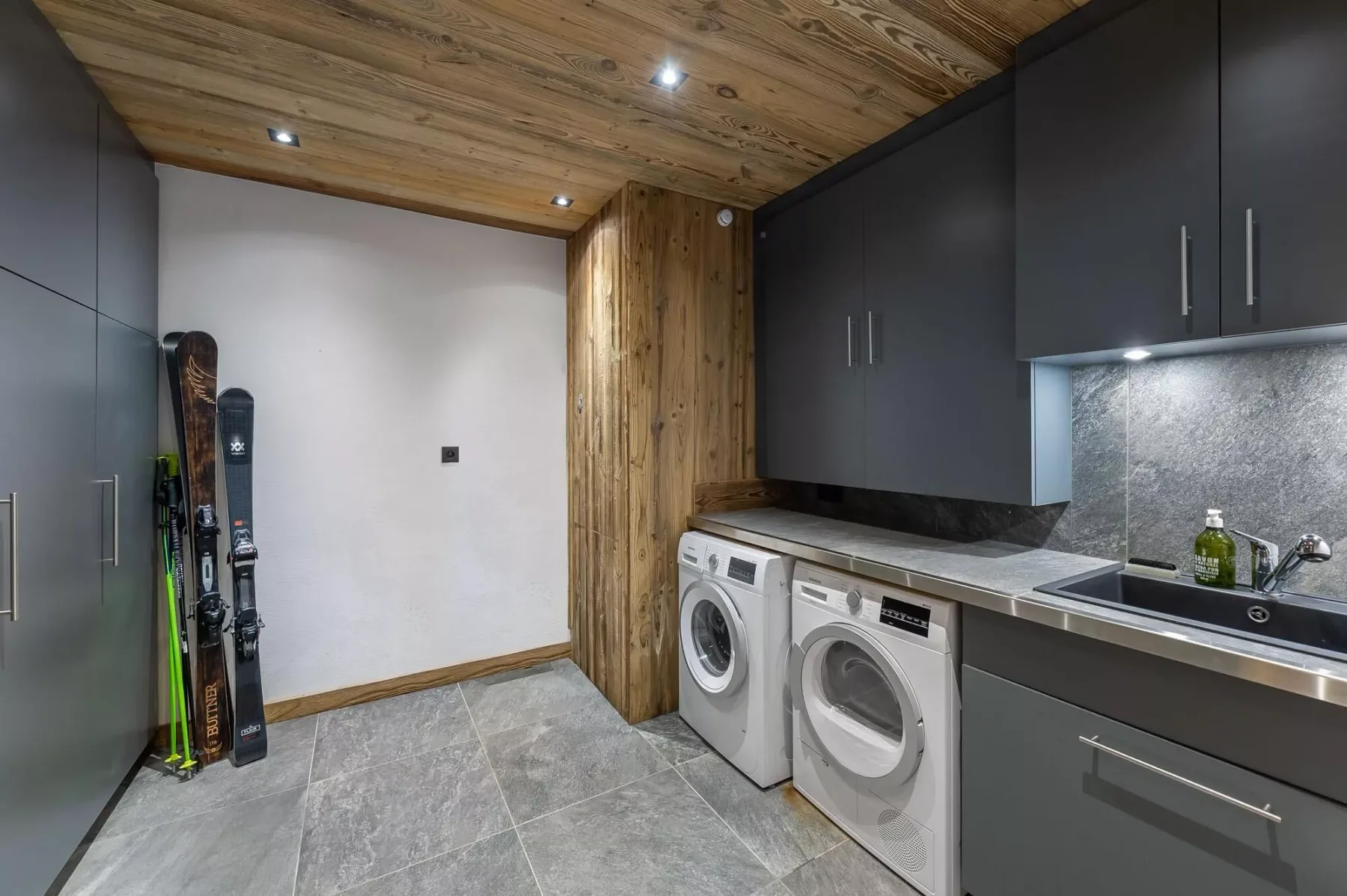 Un cuarto de lavandería con máquinas de carga frontal bajo armarios oscuros y un techo de madera.