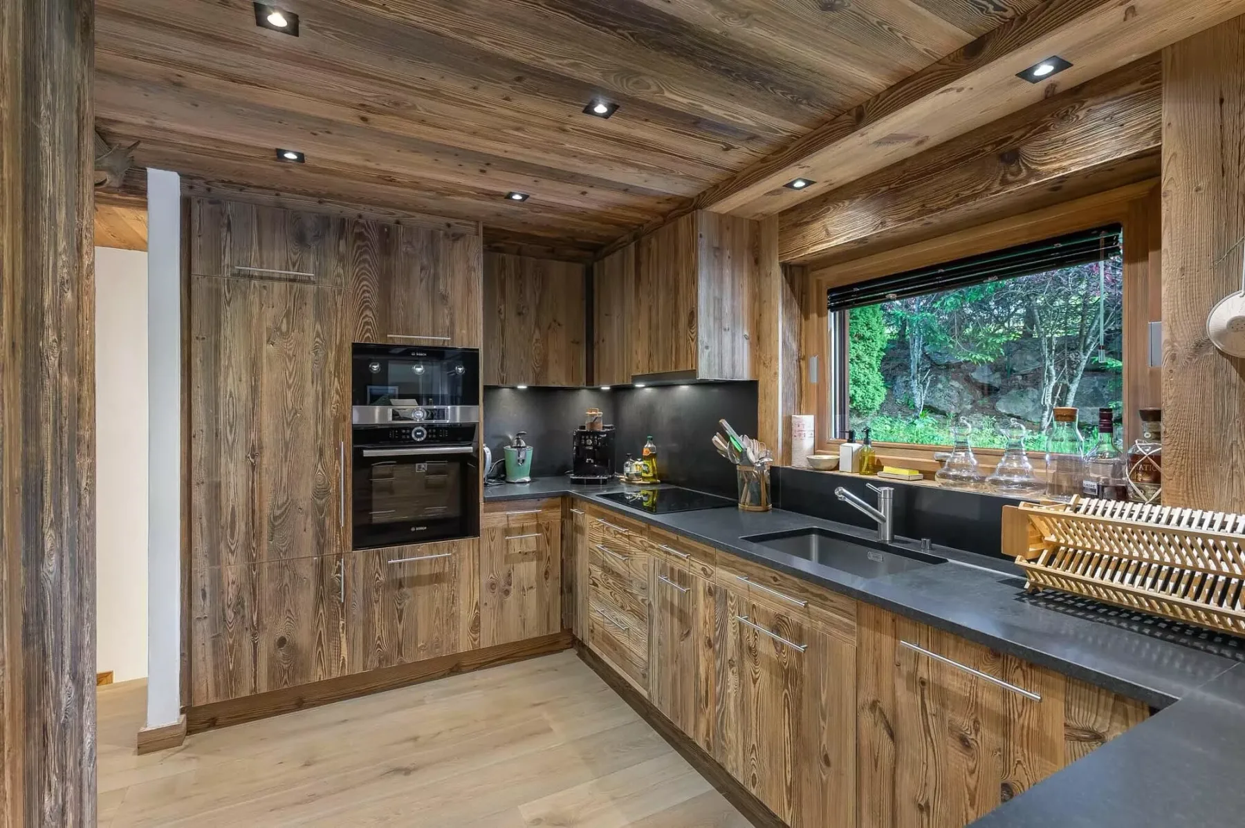 Una cocina revestida de madera, con encimeras oscuras y una ventana amplia con vistas a la vegetación.