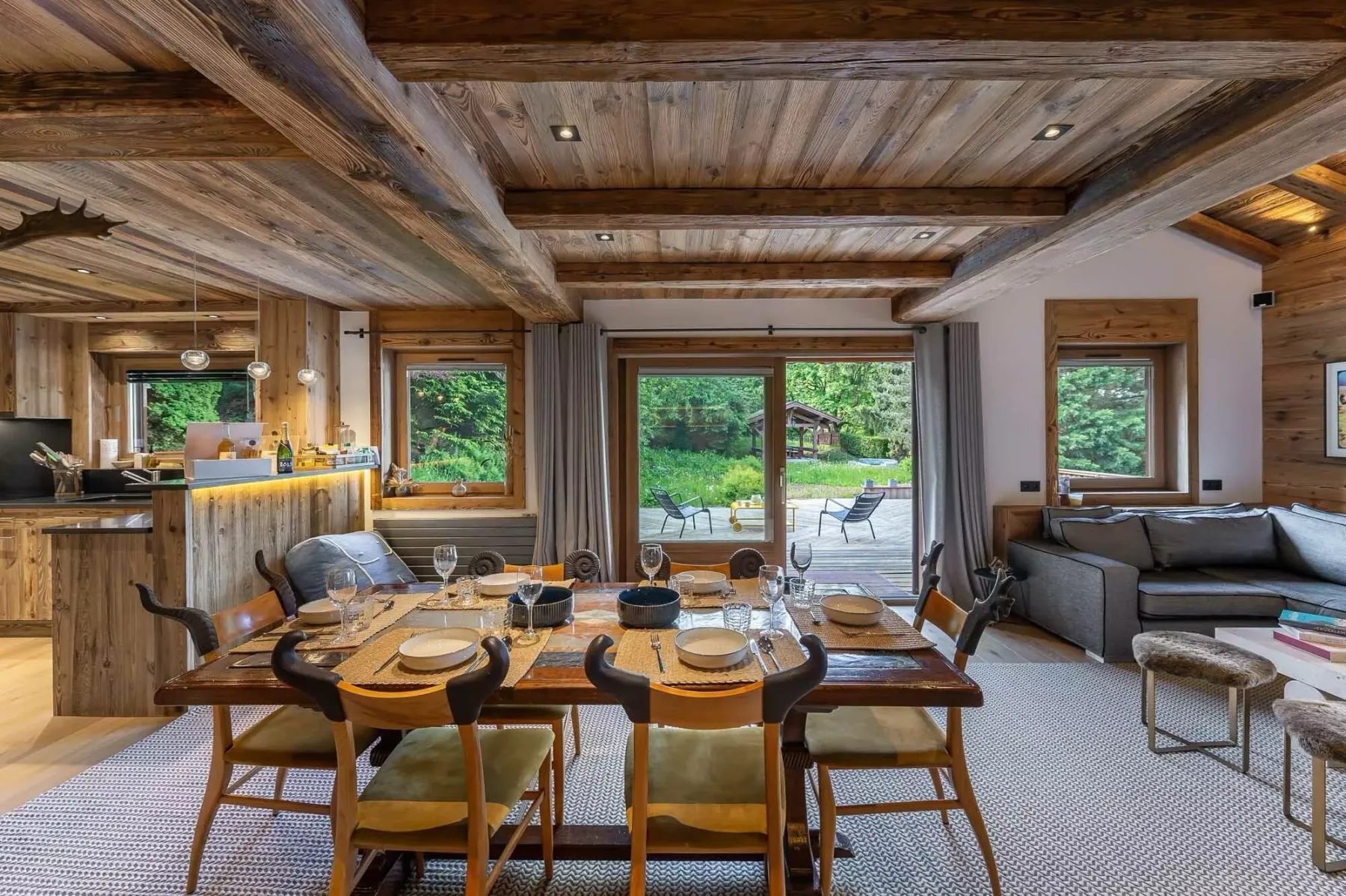 Una sala de estar y comedor de madera tiene una mesa puesta y asientos junto a grandes ventanas.
