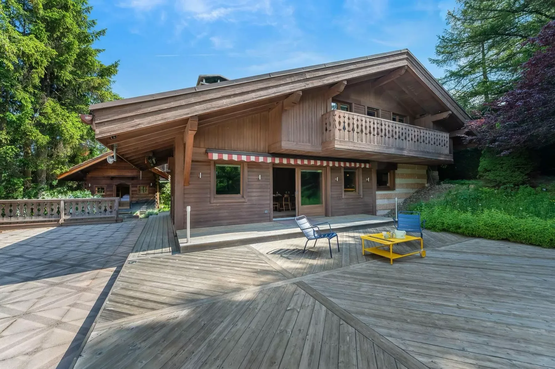 Una casa de madera con una amplia terraza, asientos al aire libre y árboles alrededor.