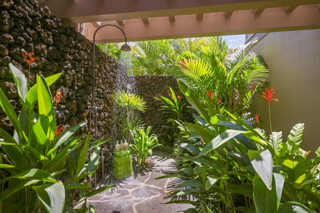 Una ducha exterior está rodeada de plantas tropicales frondosas y muros de piedra.