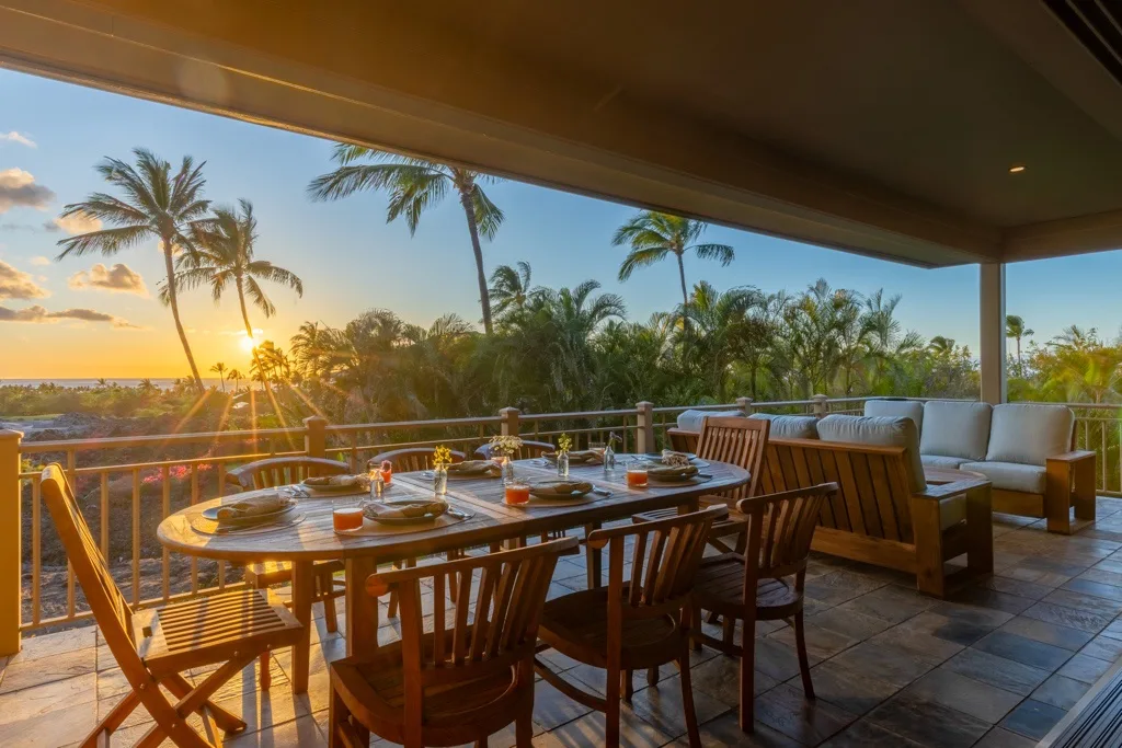 Un comedor exterior cubierto con mesa y asientos da a palmeras al atardecer.