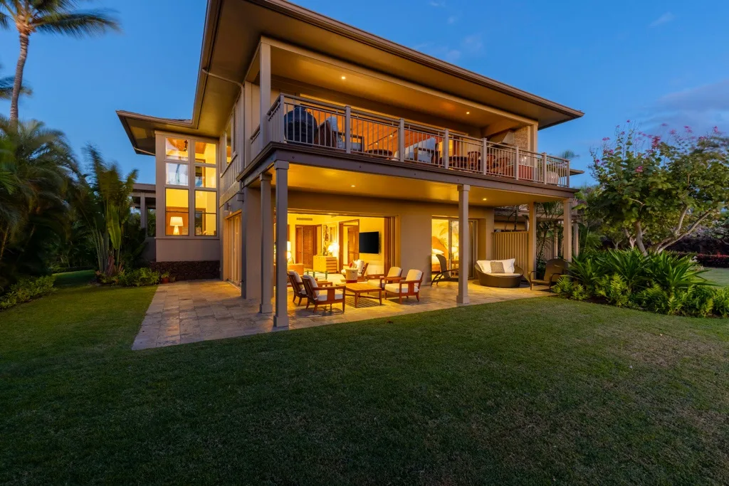 Una casa moderna con patio cubierto, balcón y césped al anochecer.