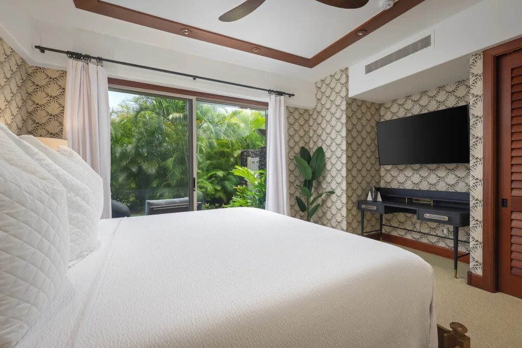 Un dormitorio con una cama blanca frente a un televisor y una ventana amplia con vegetación.