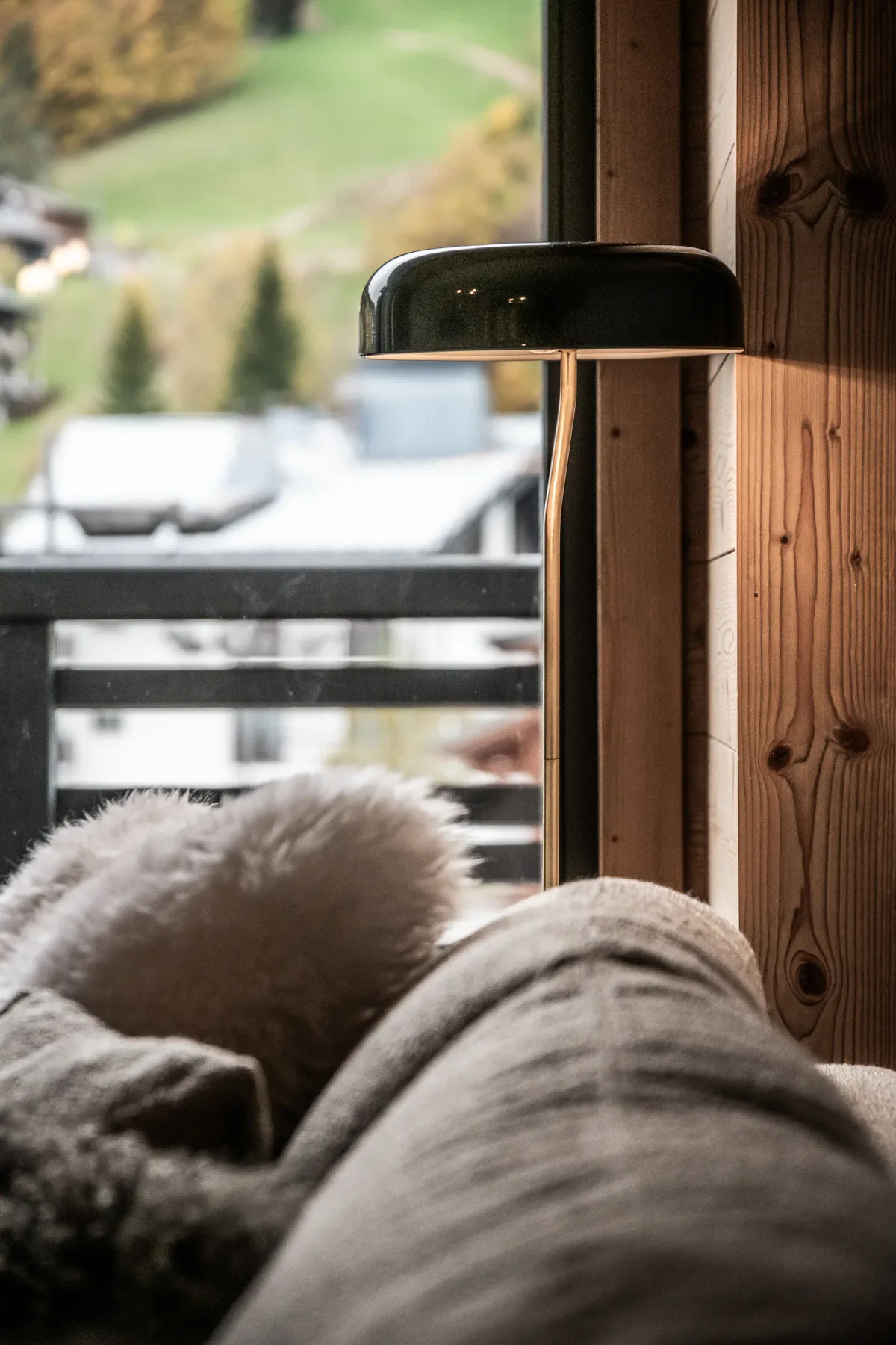 A cozy sofa with a soft blanket sits beside a wooden wall and a modern lamp, near a window overlooking a balcony and a green landscape outside.