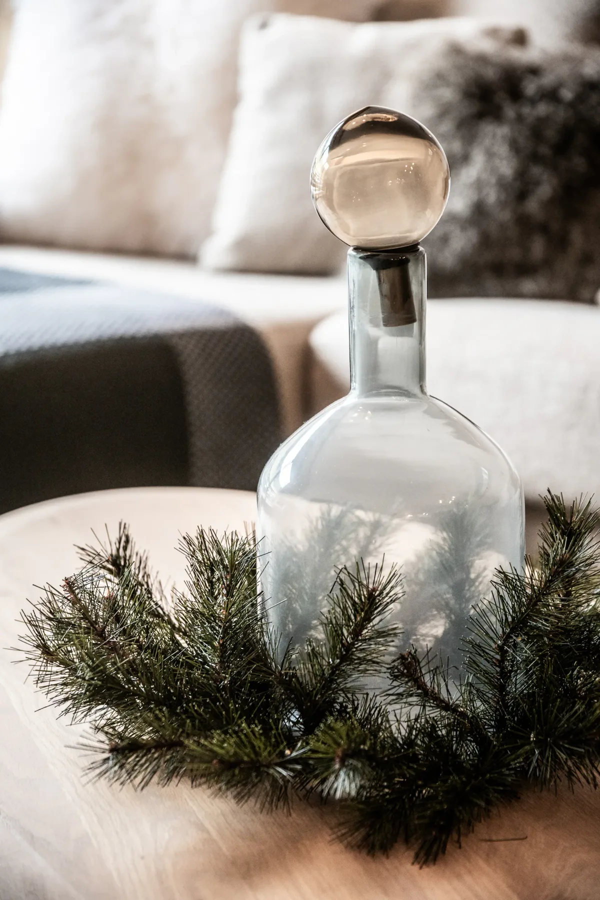 A clear glass bottle with a round stopper is placed on a soft beige surface, surrounded by a green pine wreath. A blurred couch and pillows are visible in the background.