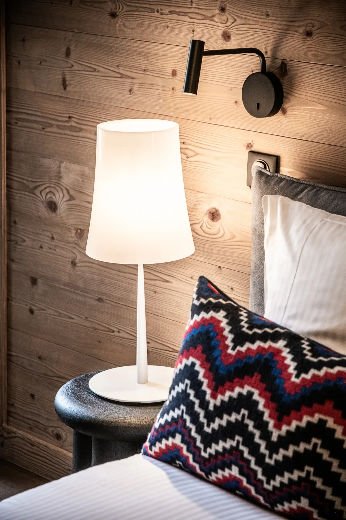 A cozy bedroom with a double bed, red and black patterned blanket, two pillows, wooden accent wall, bedside lamps, a deer artwork above the bed, red curtains, and a window with a view outside.