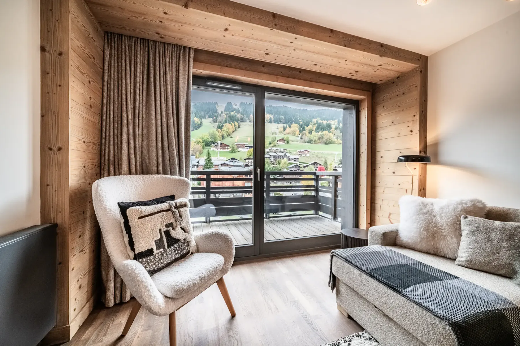 Modern living room with a large sofa, wooden coffee table, TV displaying a mountain scene, armchair, rug, and large window with a mountain view and wooden balcony outside. Neutral tones and cozy decor throughout.