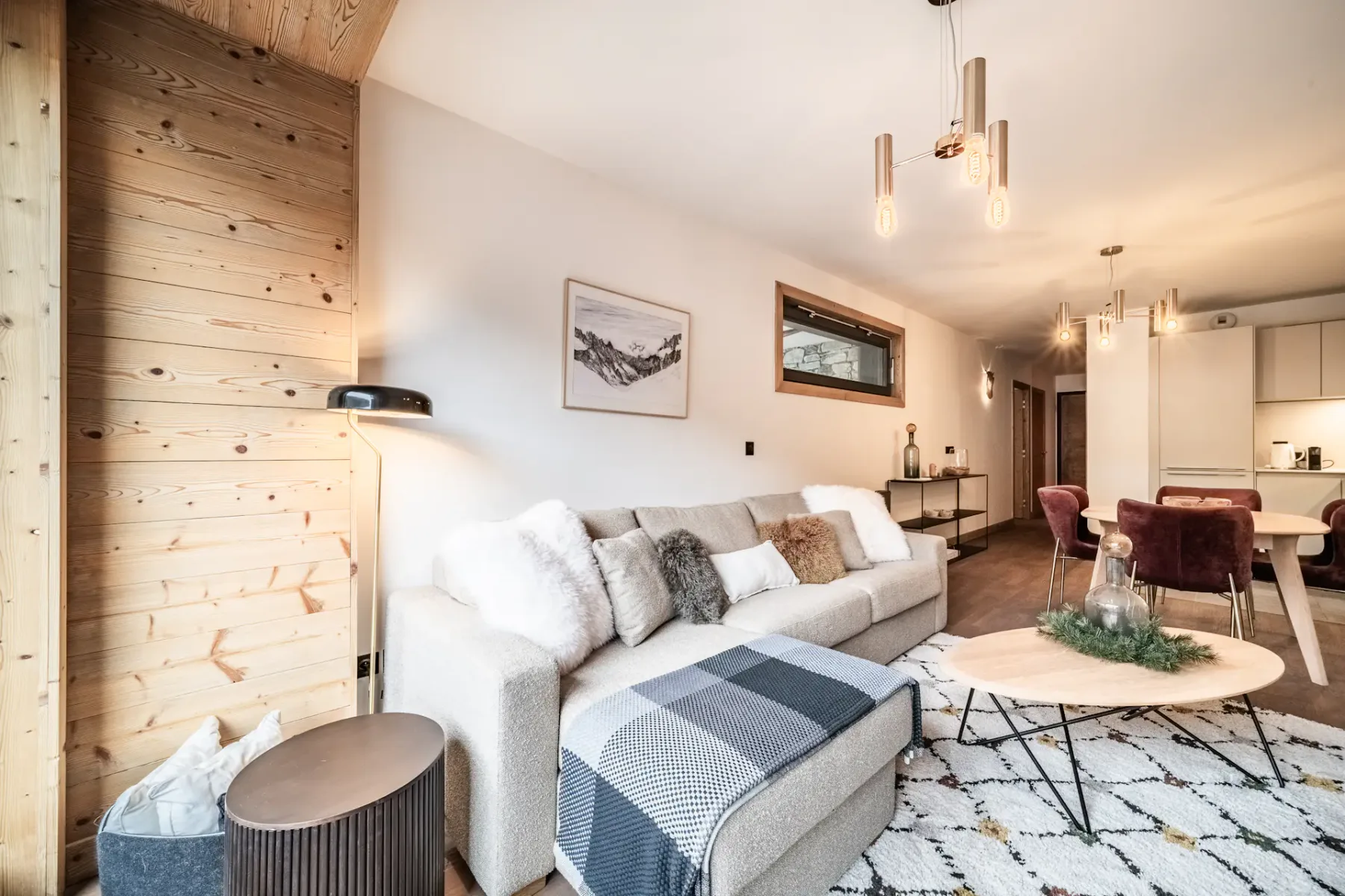 Modern living room with a large sofa, wooden coffee table, TV displaying a mountain scene, armchair, rug, and large window with a mountain view and wooden balcony outside. Neutral tones and cozy decor throughout.