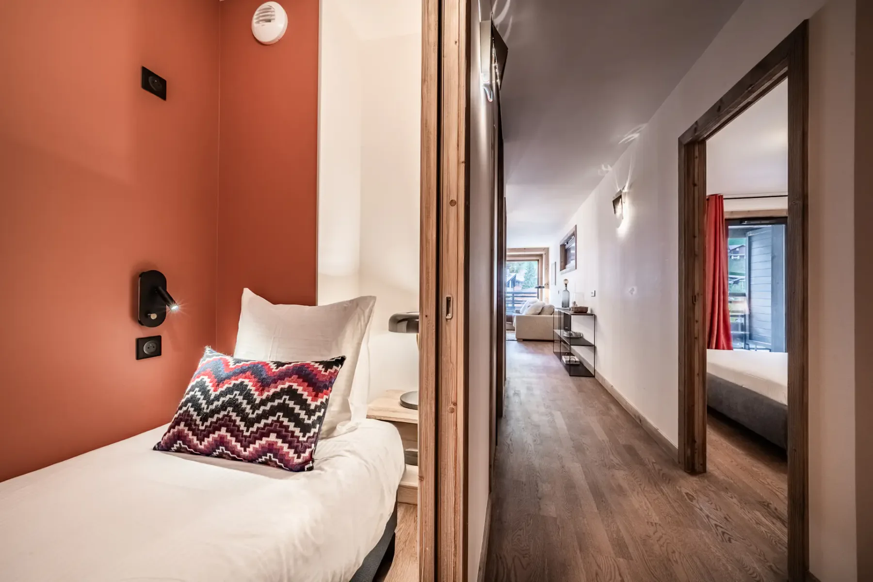 A modern hotel suite featuring a cozy single bed with a colorful zigzag pillow, a partition wall, wooden floors, and a hallway leading to a bright living area and another bedroom.