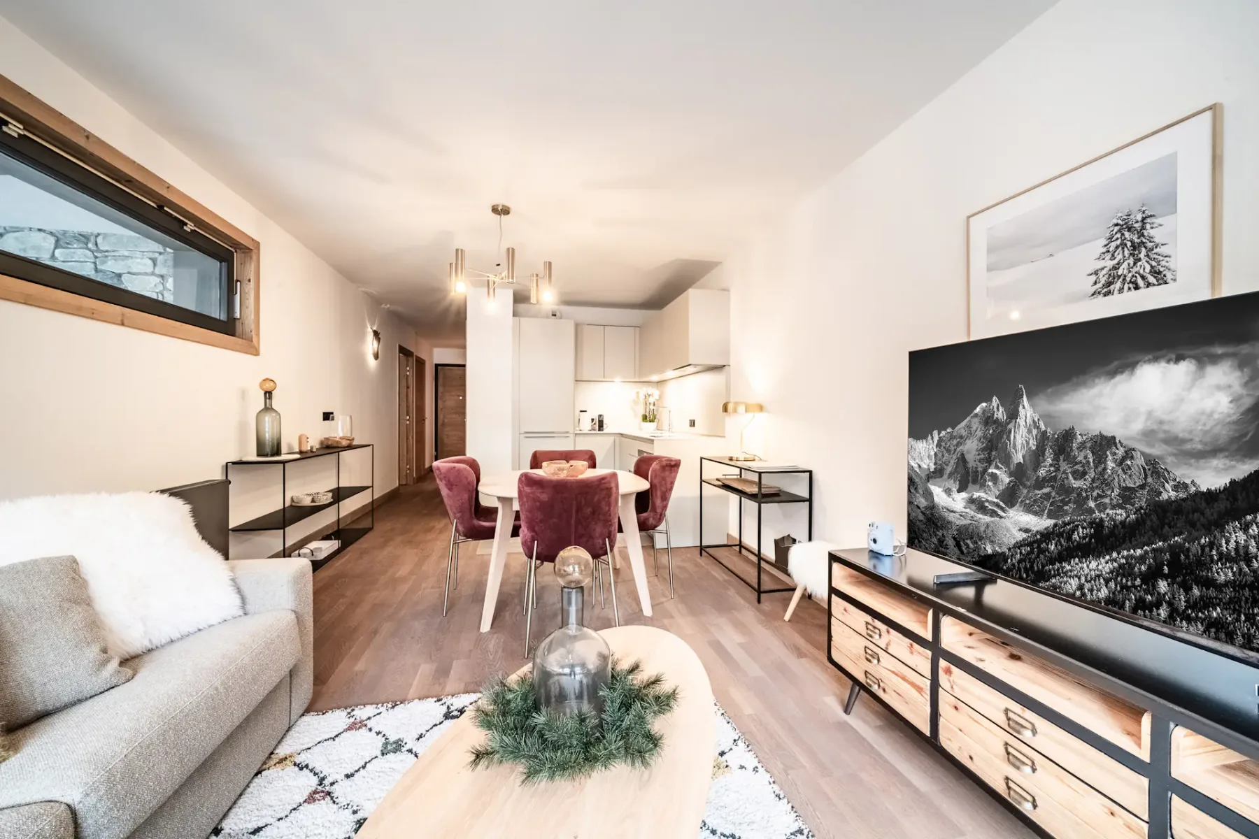 Modern living room with a large sofa, wooden coffee table, TV displaying a mountain scene, armchair, rug, and large window with a mountain view and wooden balcony outside. Neutral tones and cozy decor throughout.
