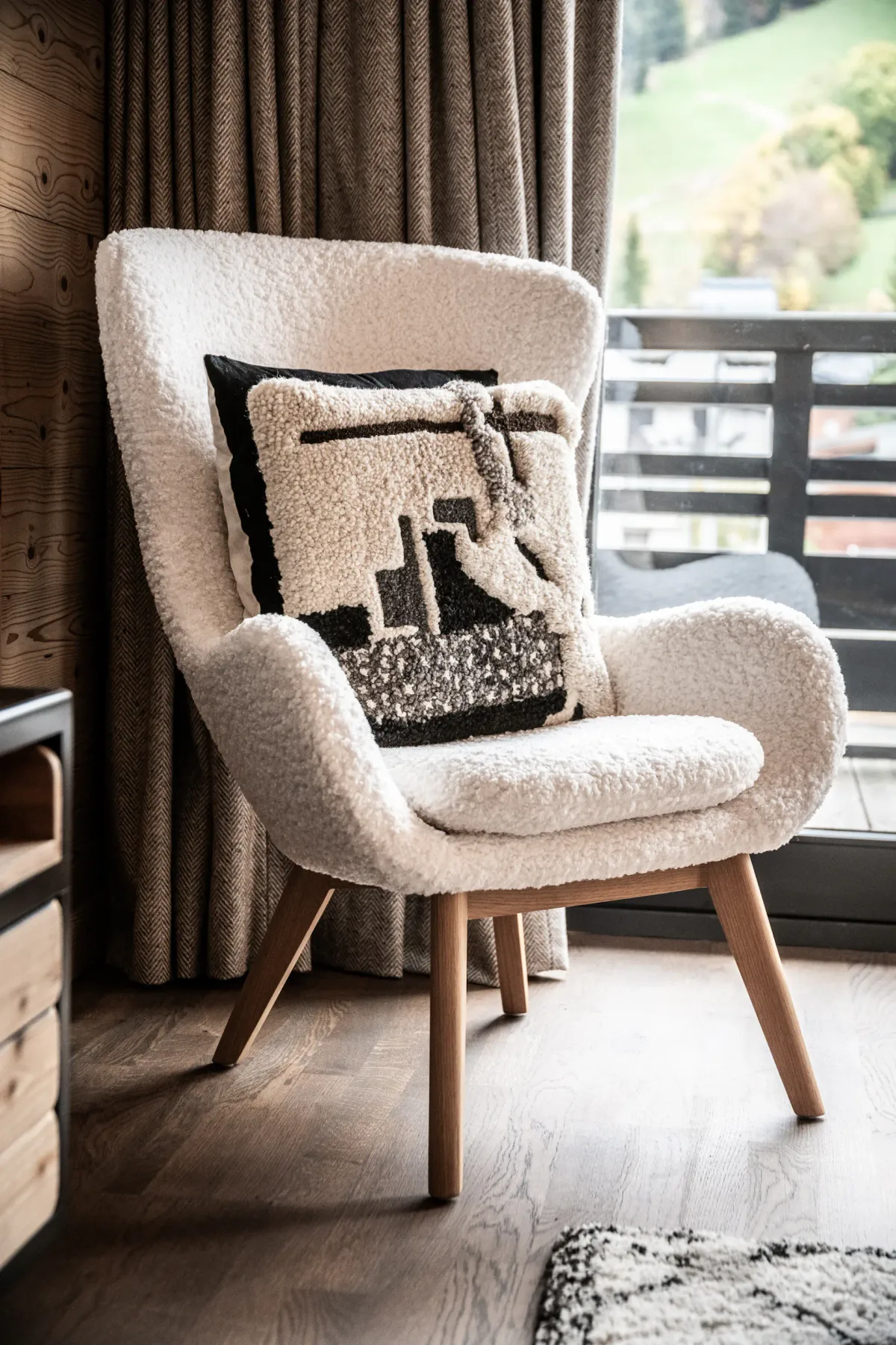 Modern living room with a large sofa, wooden coffee table, TV displaying a mountain scene, armchair, rug, and large window with a mountain view and wooden balcony outside. Neutral tones and cozy decor throughout.