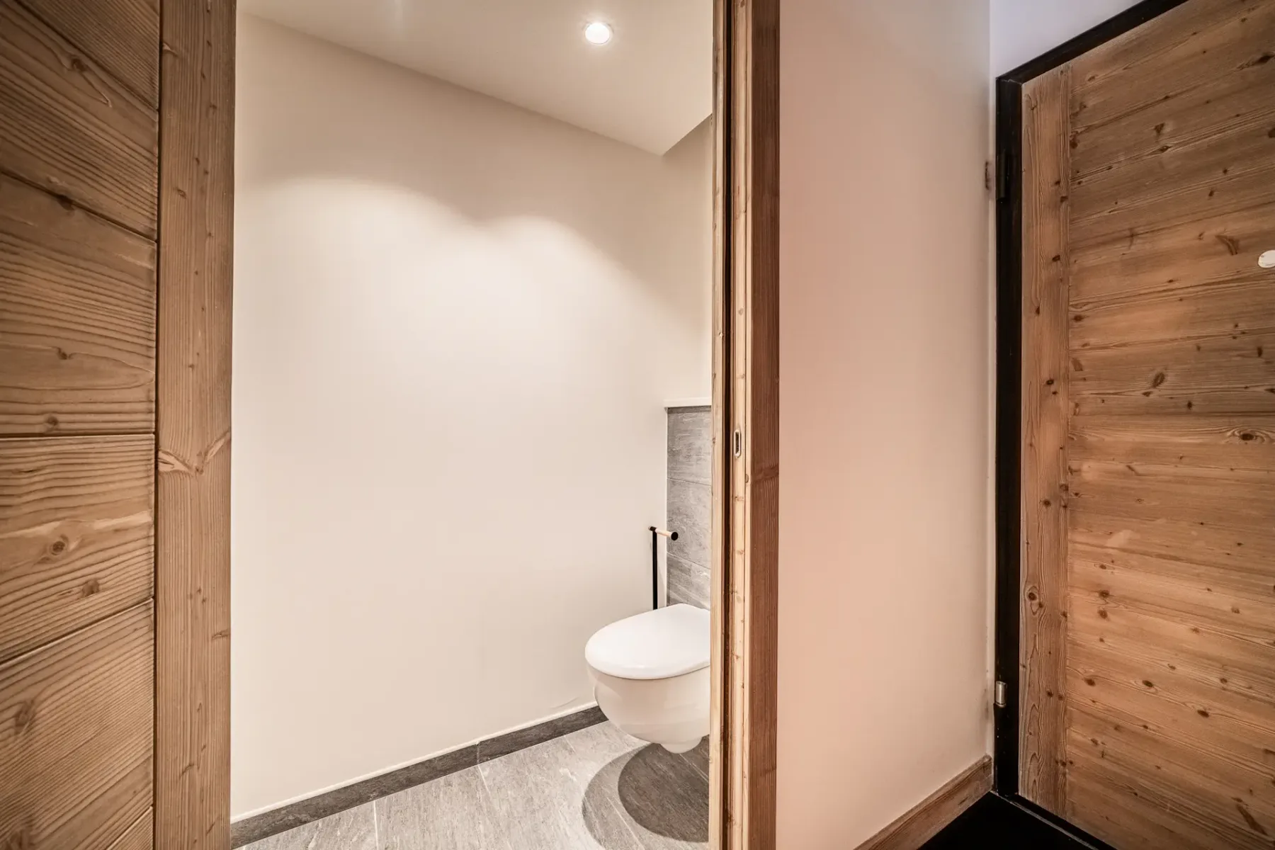 Minimalist bathroom with light-colored walls, a floating white toilet, gray tiled floor, and wood-paneled doors. The space is well-lit with recessed ceiling lights.