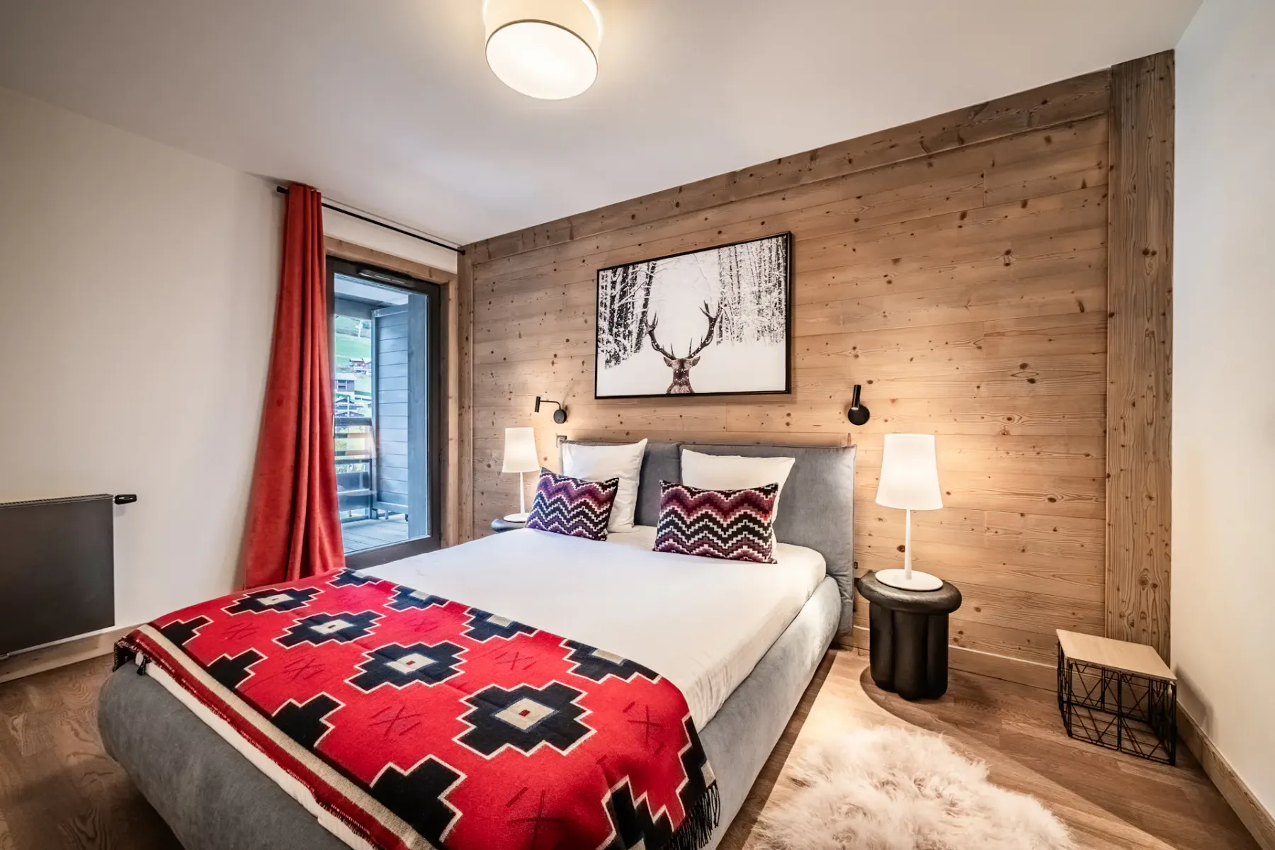 A cozy bedroom with a double bed, red and black patterned blanket, two pillows, wooden accent wall, bedside lamps, a deer artwork above the bed, red curtains, and a window with a view outside.