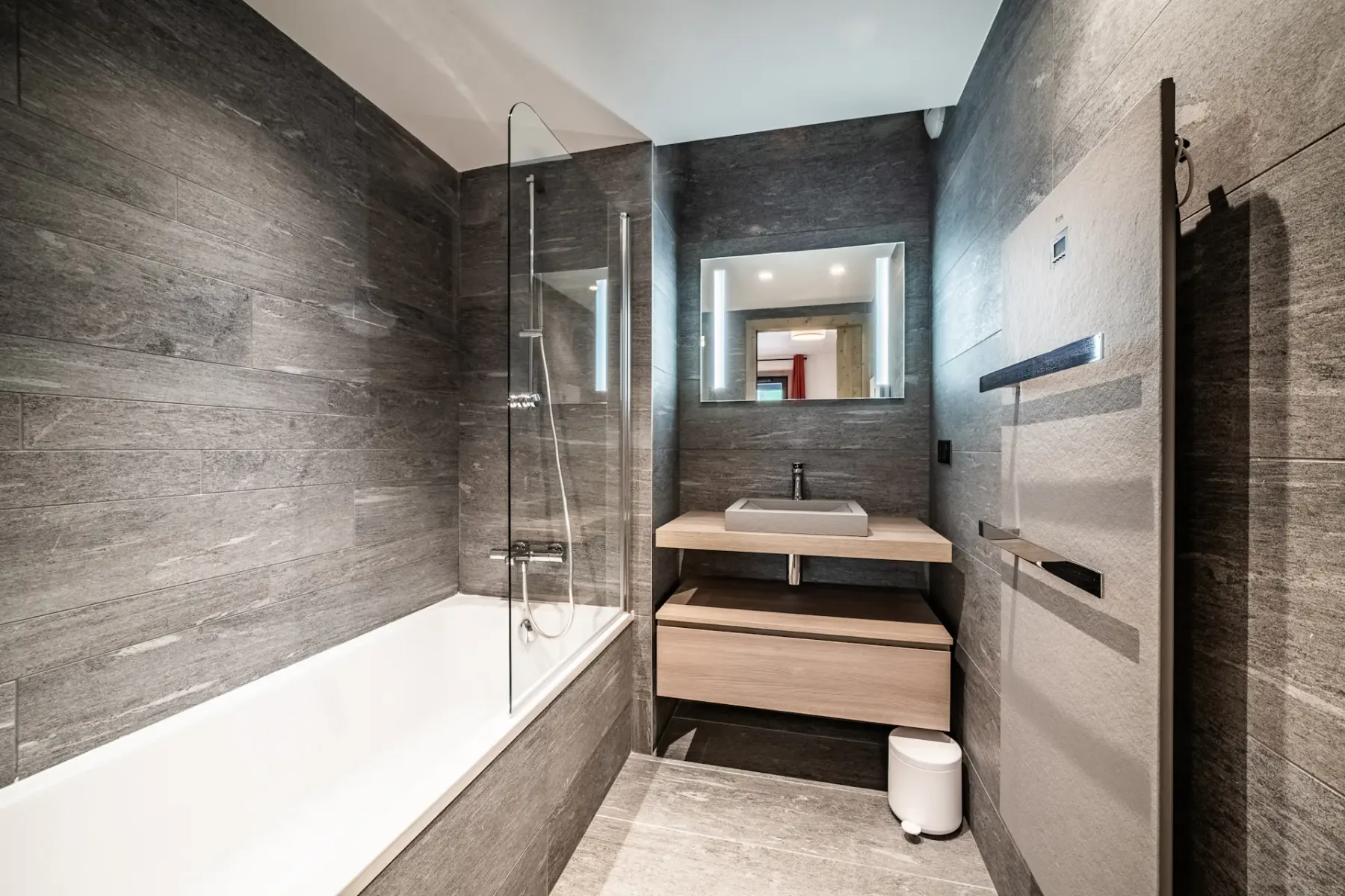 Modern bathroom with gray stone walls, a bathtub with a glass partition, a large mirror above a wooden vanity with a sink, and a small white trash bin on the floor. The space is well-lit and minimalistic.