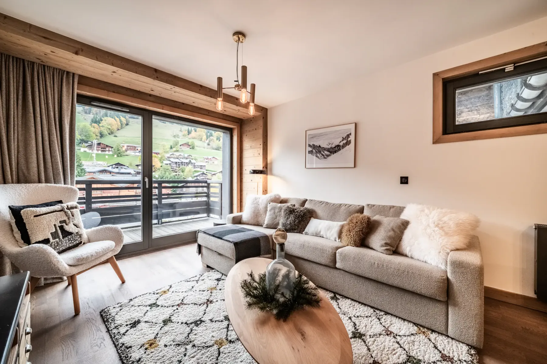 Modern living room with a large sofa, wooden coffee table, TV displaying a mountain scene, armchair, rug, and large window with a mountain view and wooden balcony outside. Neutral tones and cozy decor throughout.