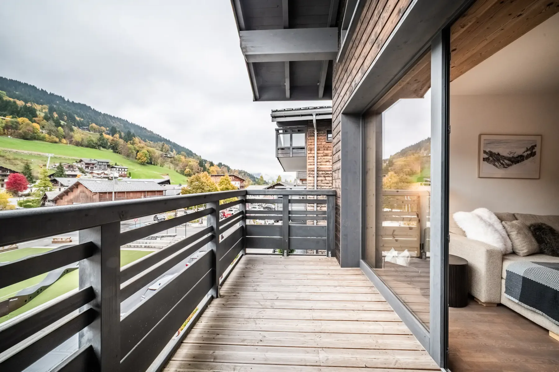 A modern wooden balcony with black railings overlooks a scenic, green hillside. Glass doors lead to a cozy living room with a sofa and airplane wall art. Overcast sky above and village buildings in the distance.
