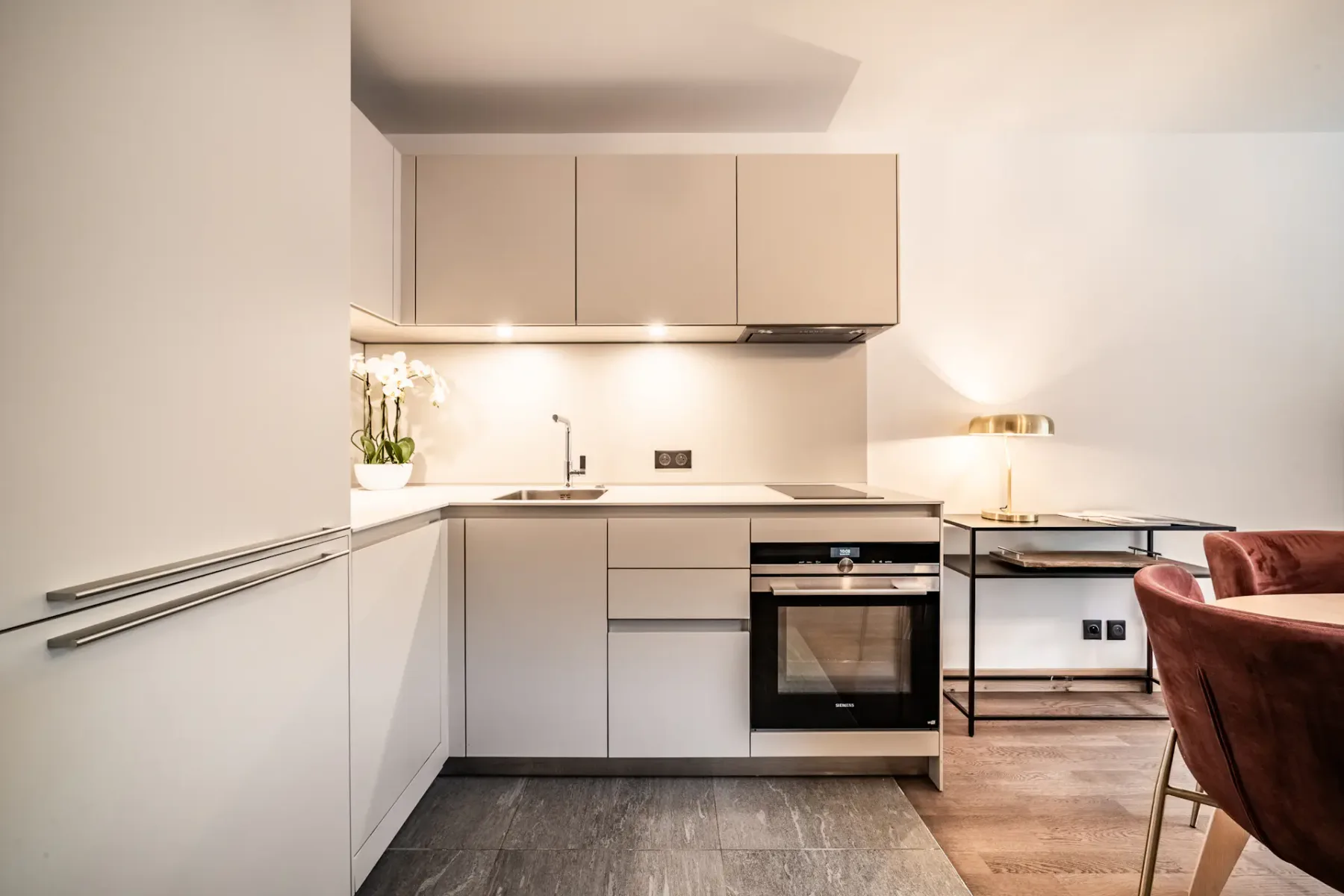 Modern, minimalist kitchen with white cabinets, built-in oven, and a small sink. There is a table with red chairs and a lamp in the background, with soft lighting creating a cozy atmosphere.