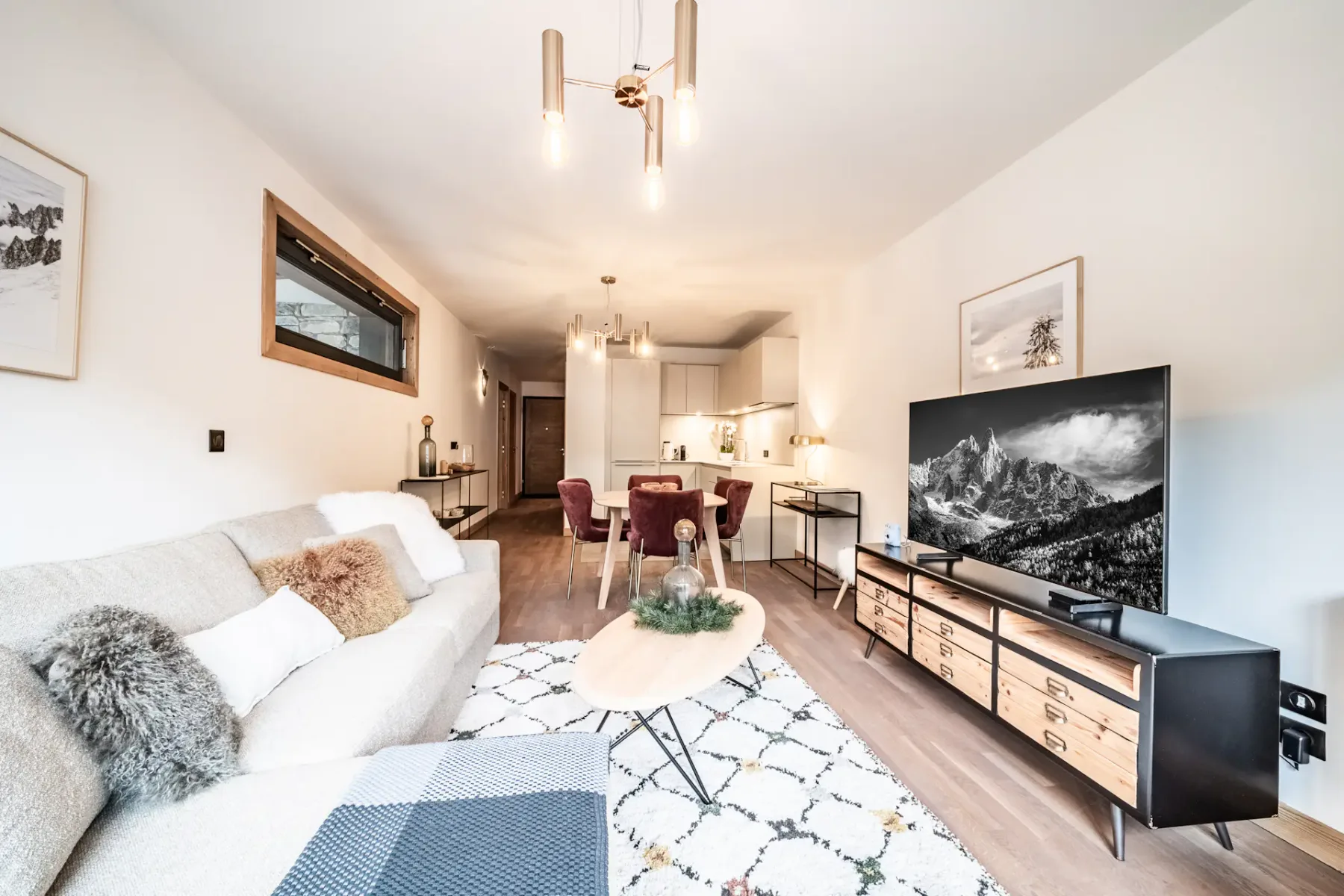 Modern living room with a large sofa, wooden coffee table, TV displaying a mountain scene, armchair, rug, and large window with a mountain view and wooden balcony outside. Neutral tones and cozy decor throughout.