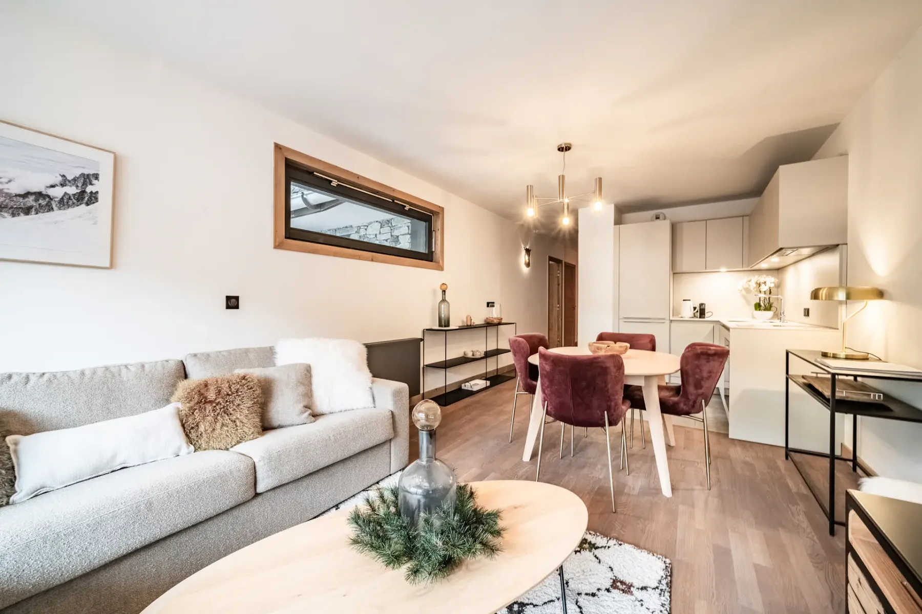 Modern living room with a large sofa, wooden coffee table, TV displaying a mountain scene, armchair, rug, and large window with a mountain view and wooden balcony outside. Neutral tones and cozy decor throughout.