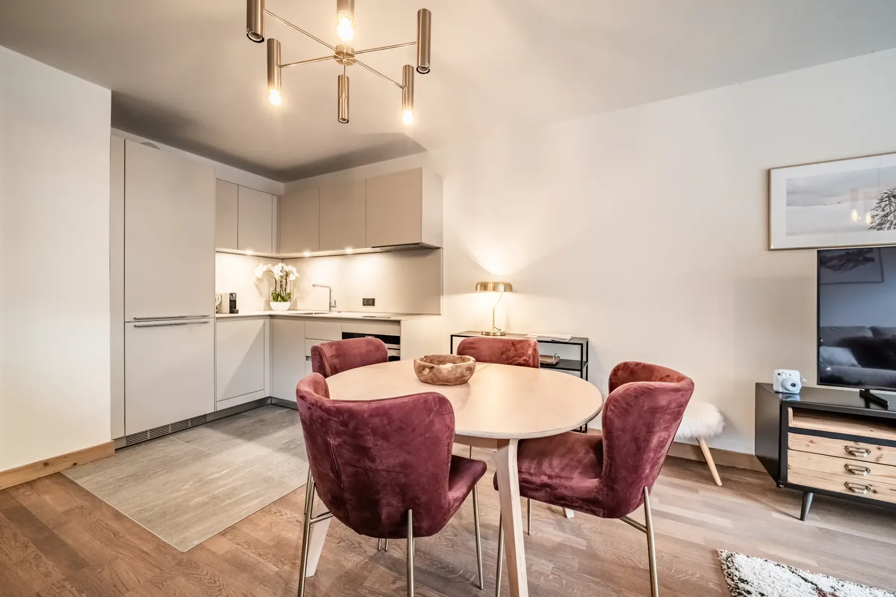 Modern, minimalist kitchen with white cabinets, built-in oven, and a small sink. There is a table with red chairs and a lamp in the background, with soft lighting creating a cozy atmosphere.