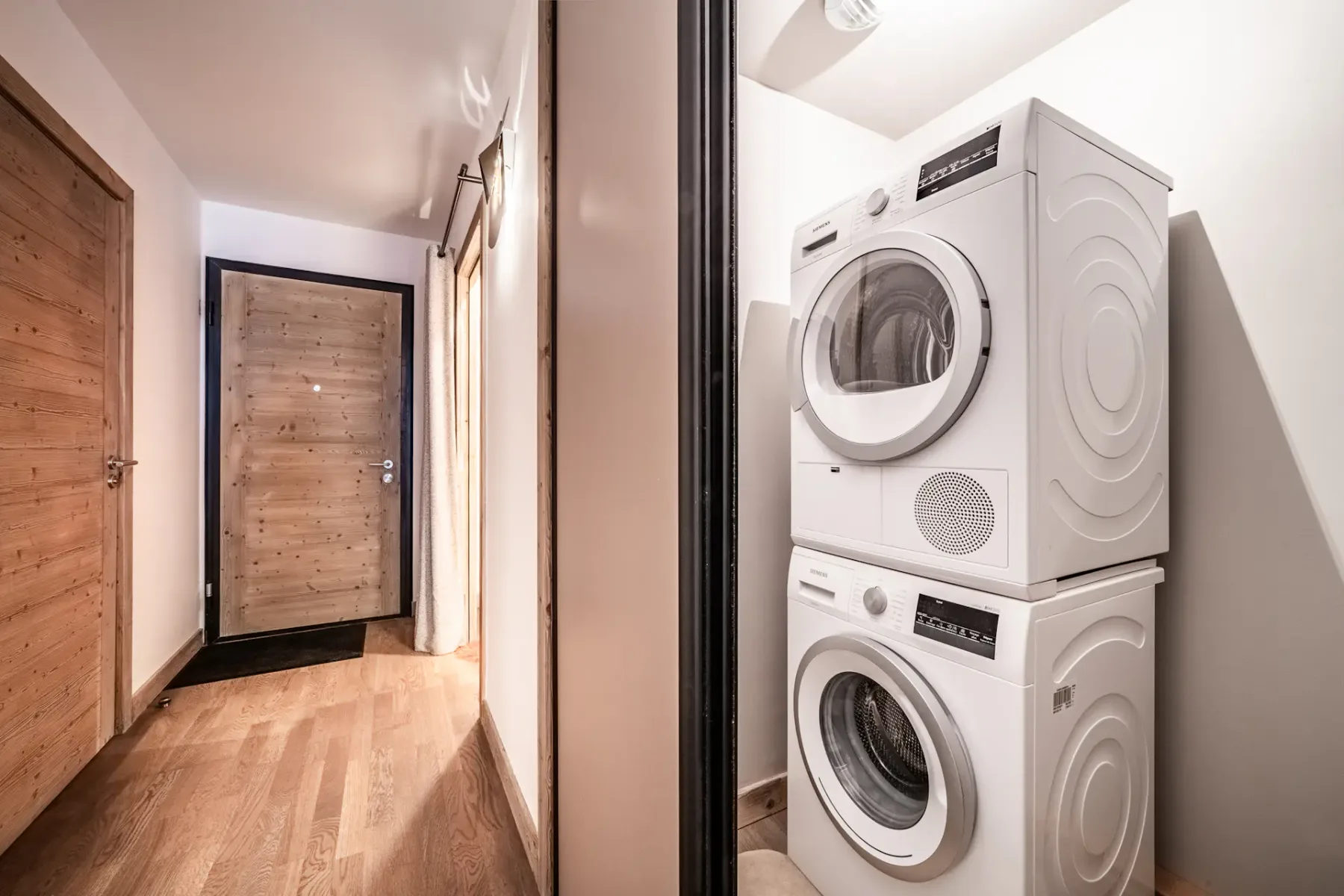 A modern laundry nook with a stacked white washer and dryer is located in a small alcove off a hallway with wooden floors and doors. The area is well-lit and organized.