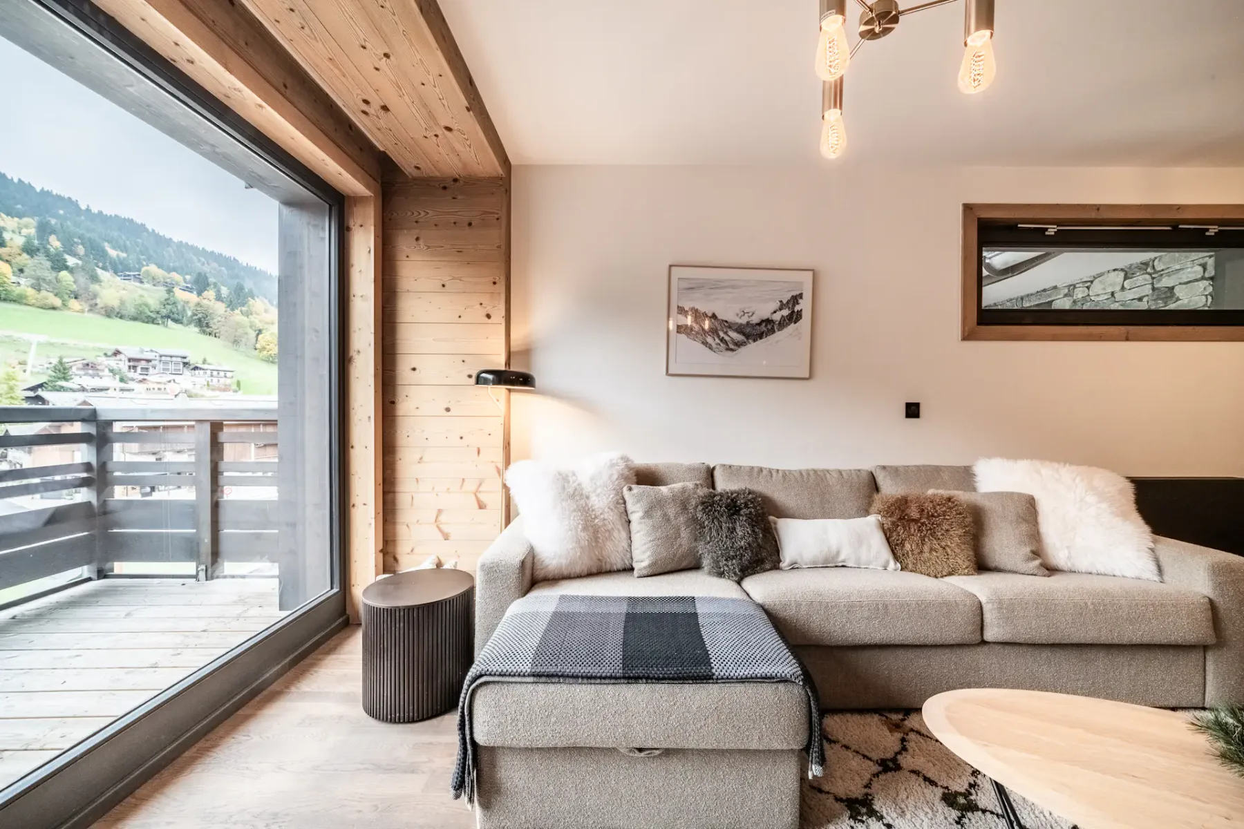 Modern living room with a large sofa, wooden coffee table, TV displaying a mountain scene, armchair, rug, and large window with a mountain view and wooden balcony outside. Neutral tones and cozy decor throughout.