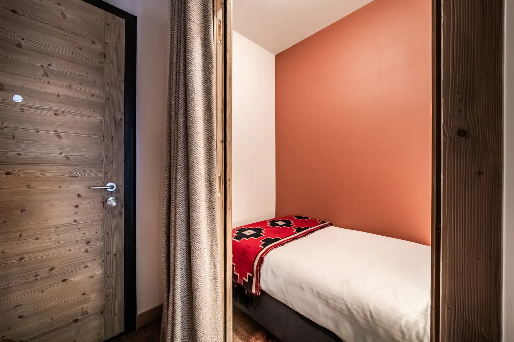 A modern hotel suite featuring a cozy single bed with a colorful zigzag pillow, a partition wall, wooden floors, and a hallway leading to a bright living area and another bedroom.