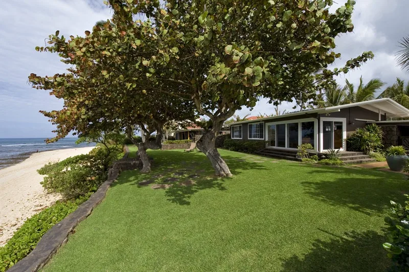 A beachfront house stands beside a grassy lawn shaded by leafy trees.
