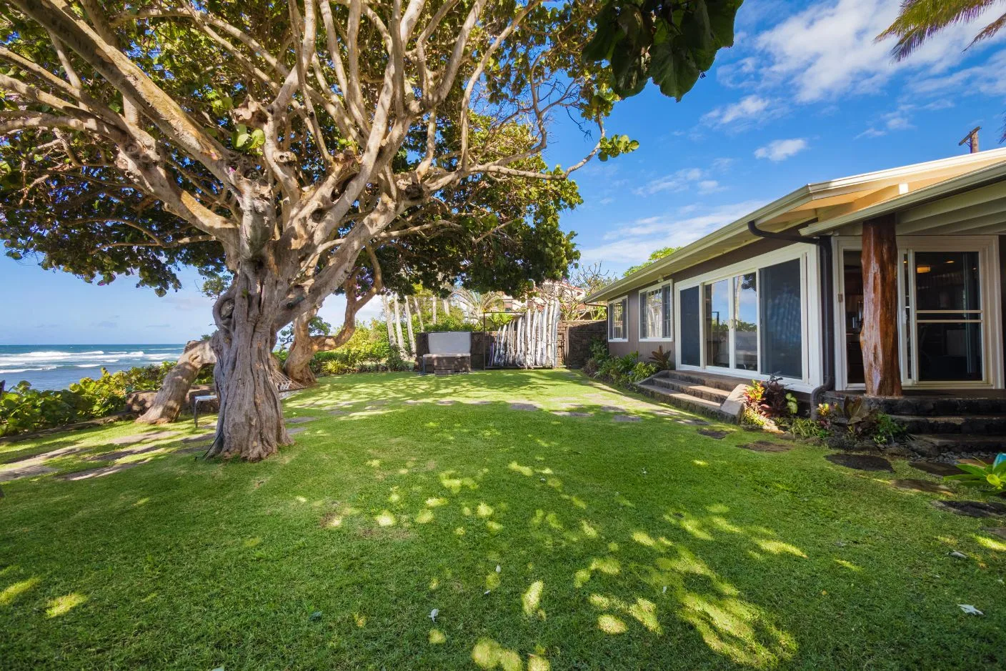 A grassy yard beside a low house, with a large leafy tree and ocean views.