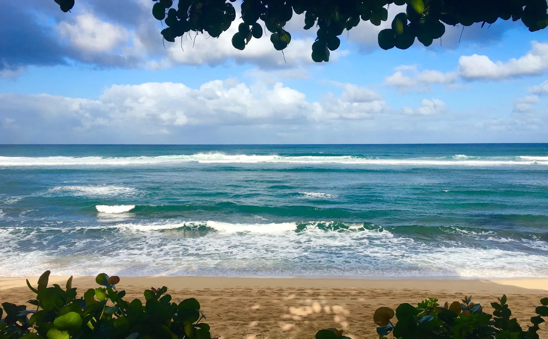 A sandy beach faces rolling ocean waves beneath a partly cloudy sky.