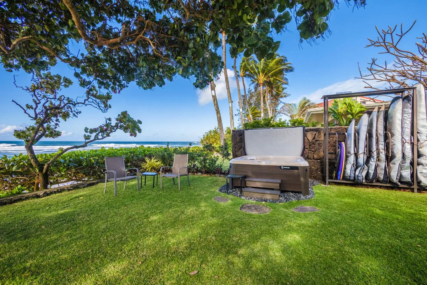 A grassy seaside garden with outdoor seating, a hot tub, and surfboards.