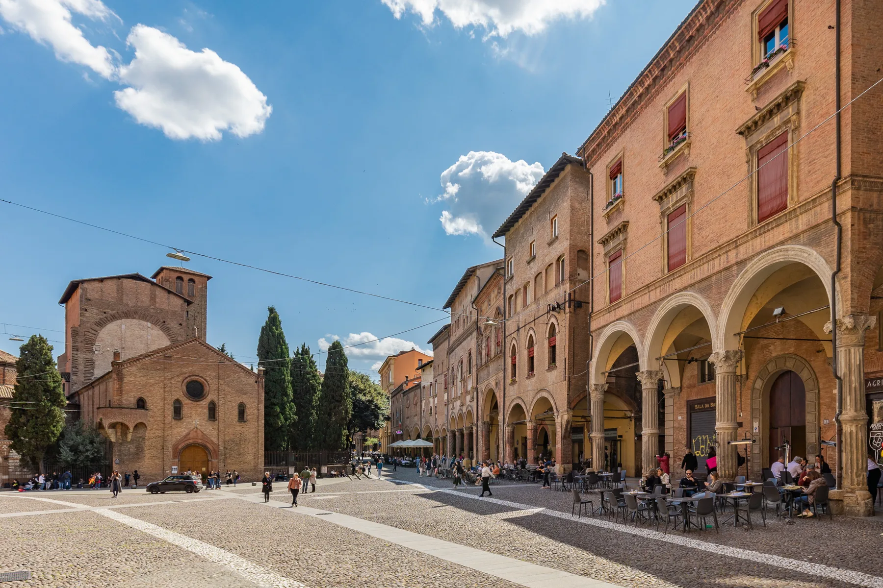 A sunlit town square with historic buildings and arcades beneath a blue sky.