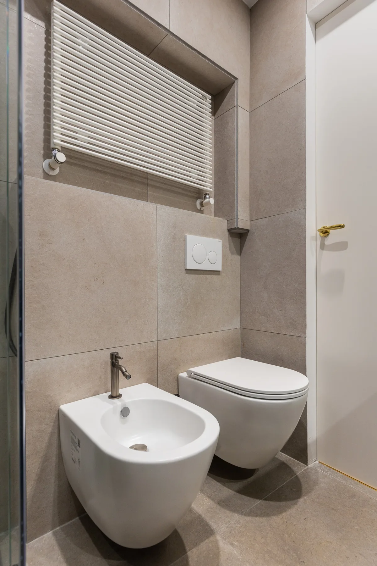 A tiled bathroom with a bidet and toilet beneath a window blind.