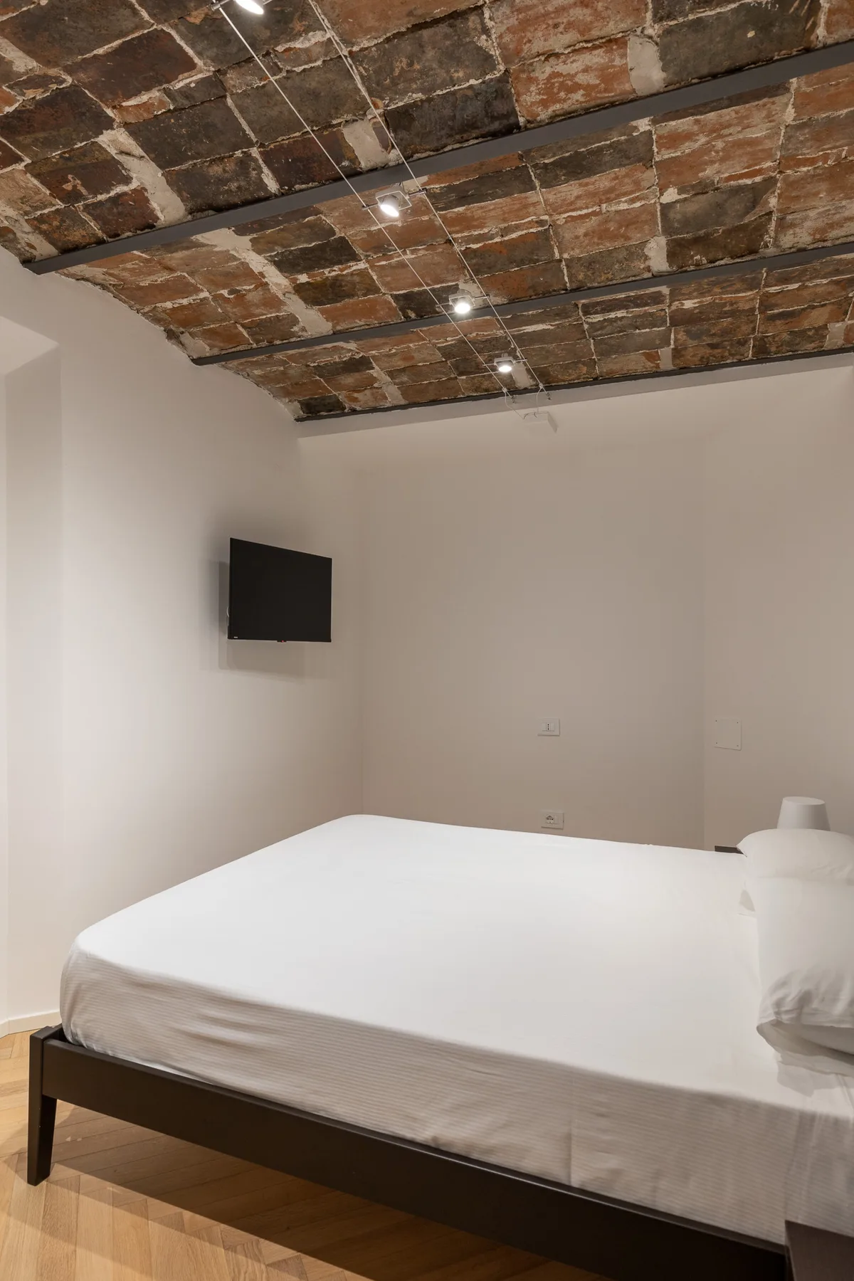 A bedroom with a large bed, wall-mounted television, and exposed brick ceiling.