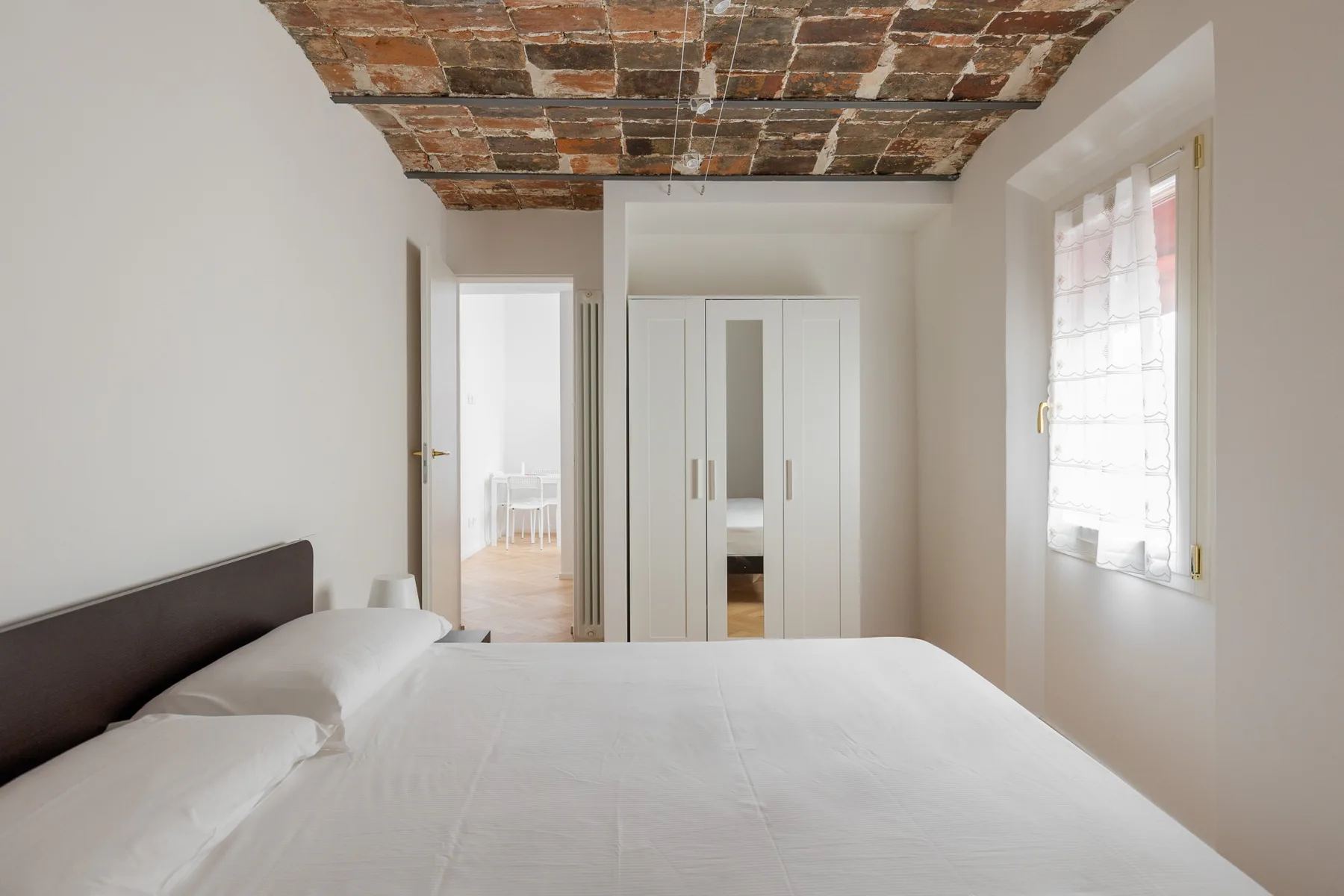 A bedroom with a neatly made bed, a wardrobe, and an exposed brick ceiling.