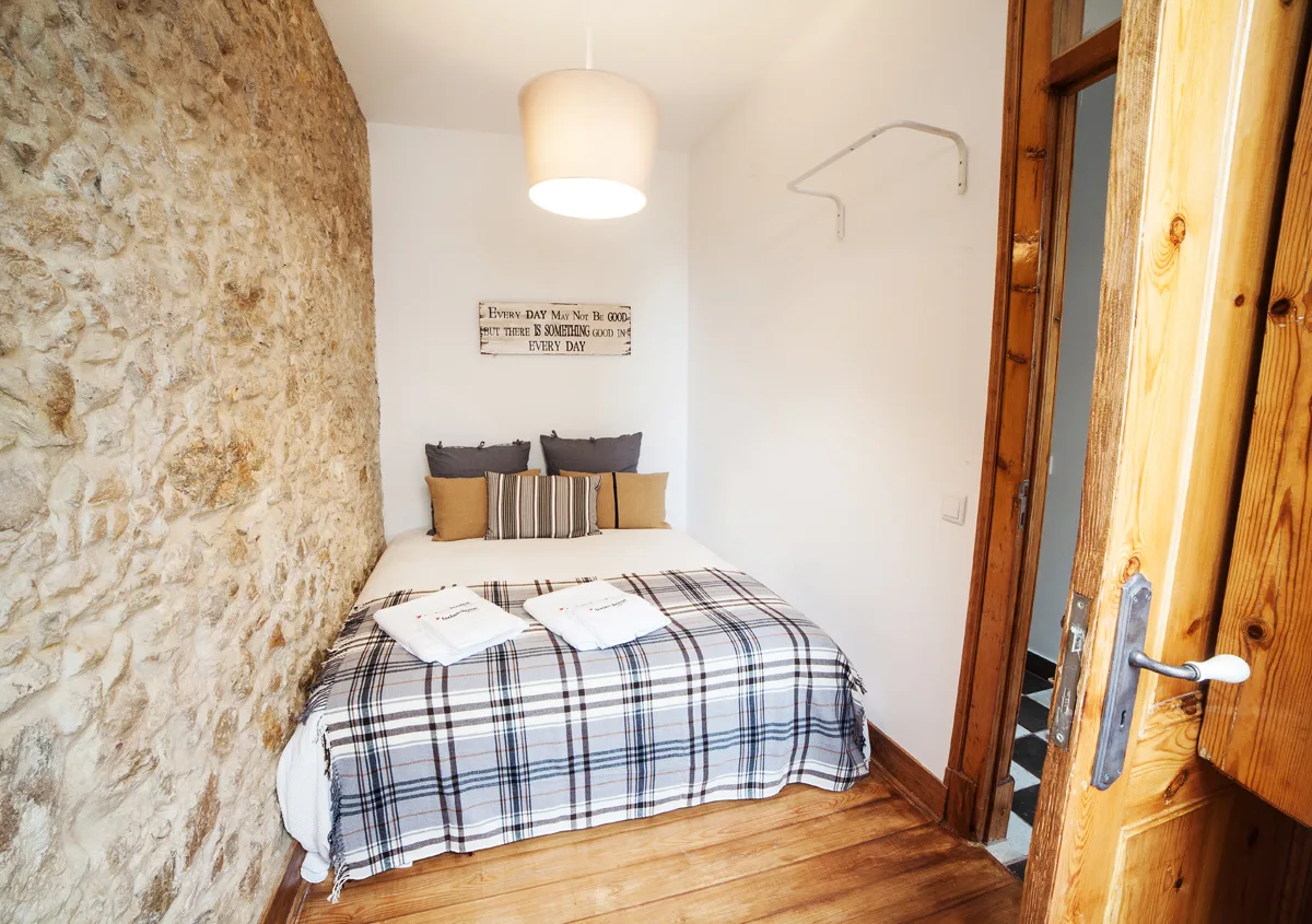 A compact bedroom with a plaid-covered bed beside a stone wall.