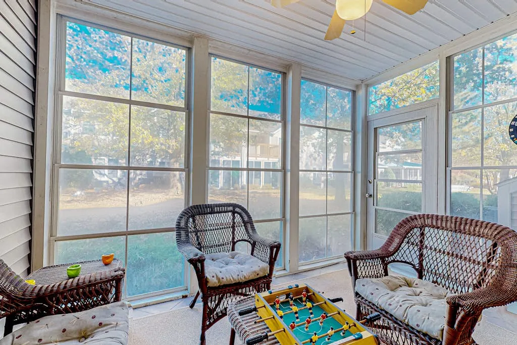 A bright sunroom with large windows and wicker seating around a small game table.