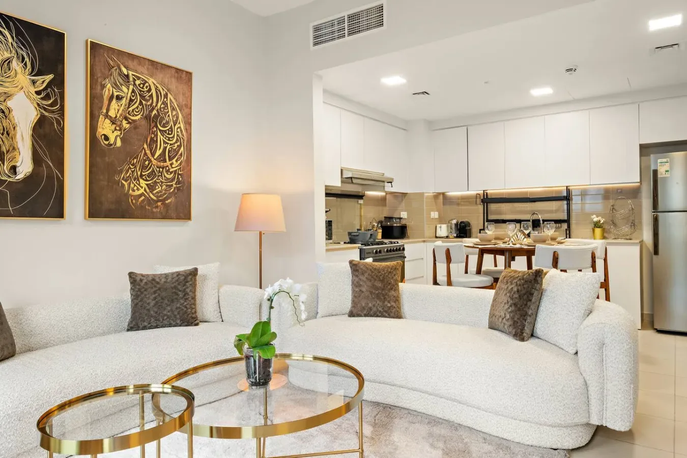 A bright living room with curved seating, glass tables, and an open kitchen.