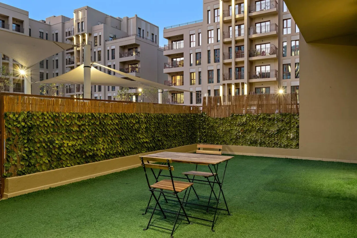 An outdoor seating area with a small table sits beside a green hedge.