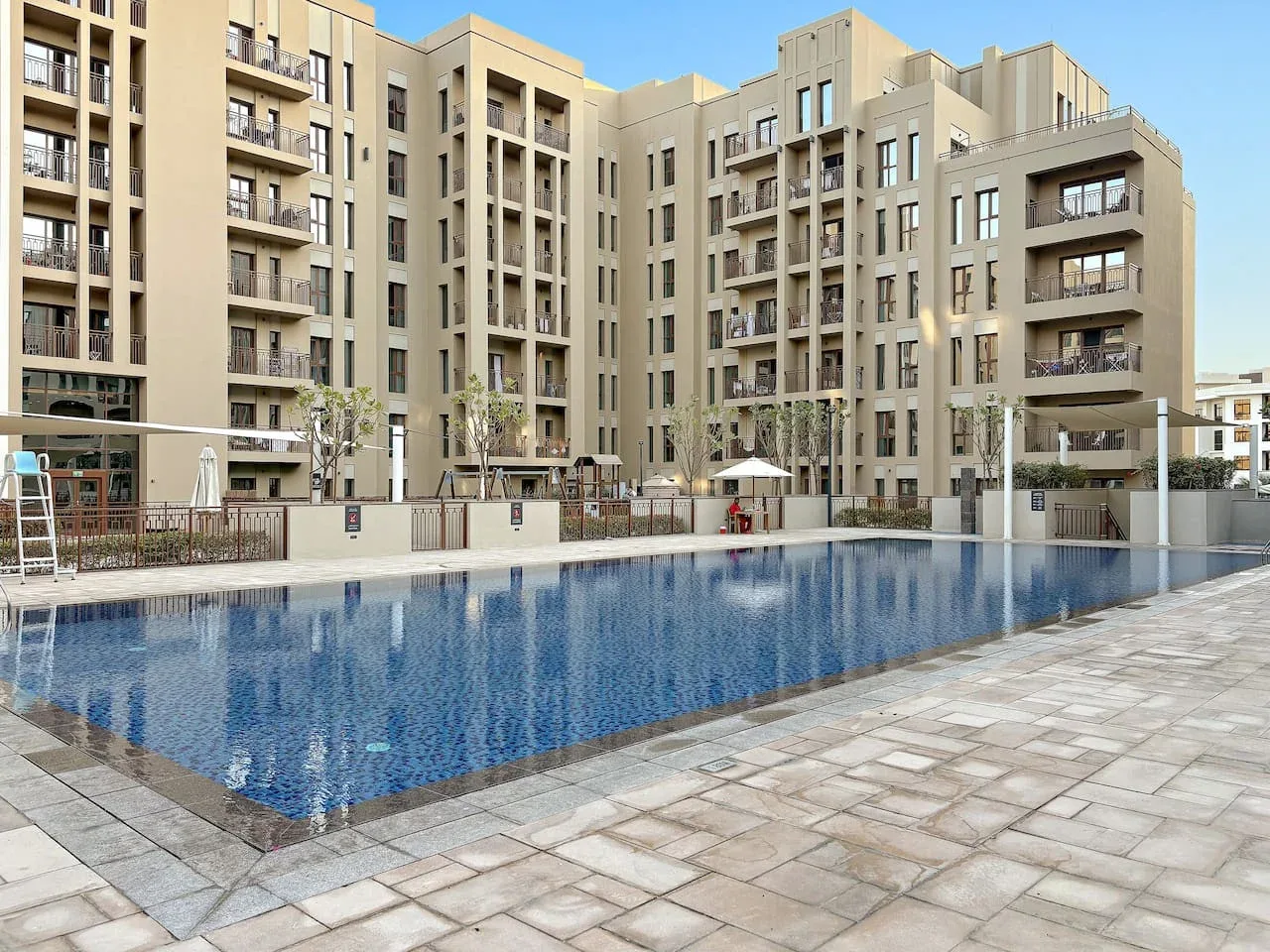 An outdoor swimming pool sits beside a paved deck in front of beige apartment buildings.