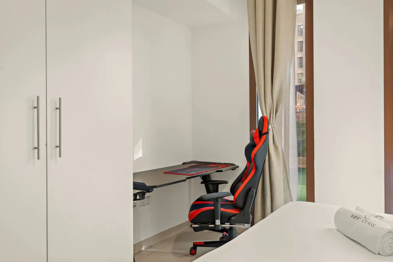 A bedroom with a gaming chair and desk beside a window.