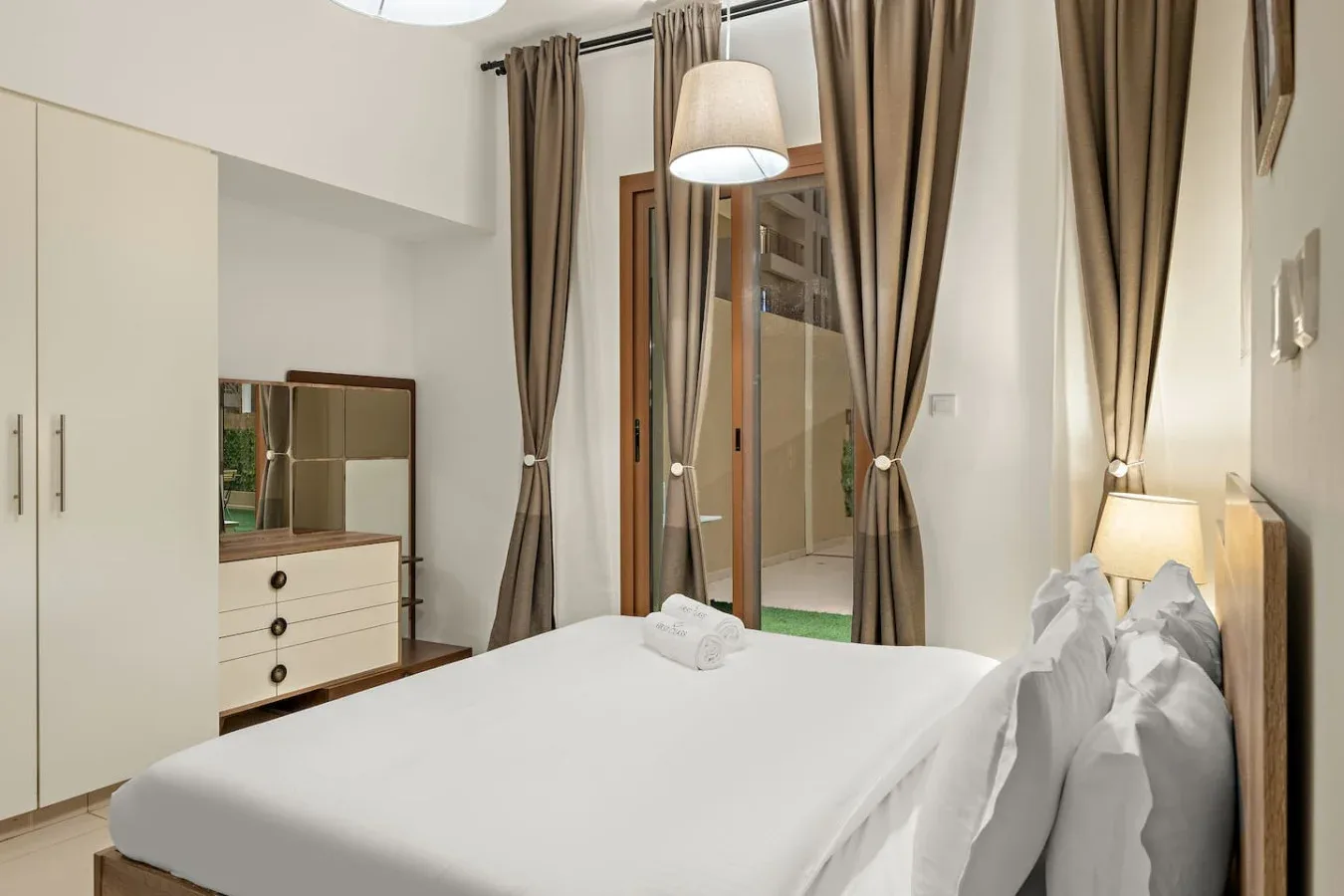 A bedroom with a neatly made bed, a dresser, and curtains by the glass doors.