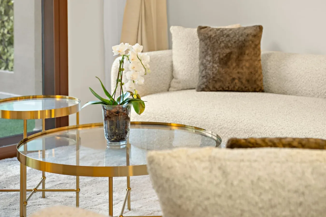 A bright living room with cream seating, glass tables, and a potted orchid.
