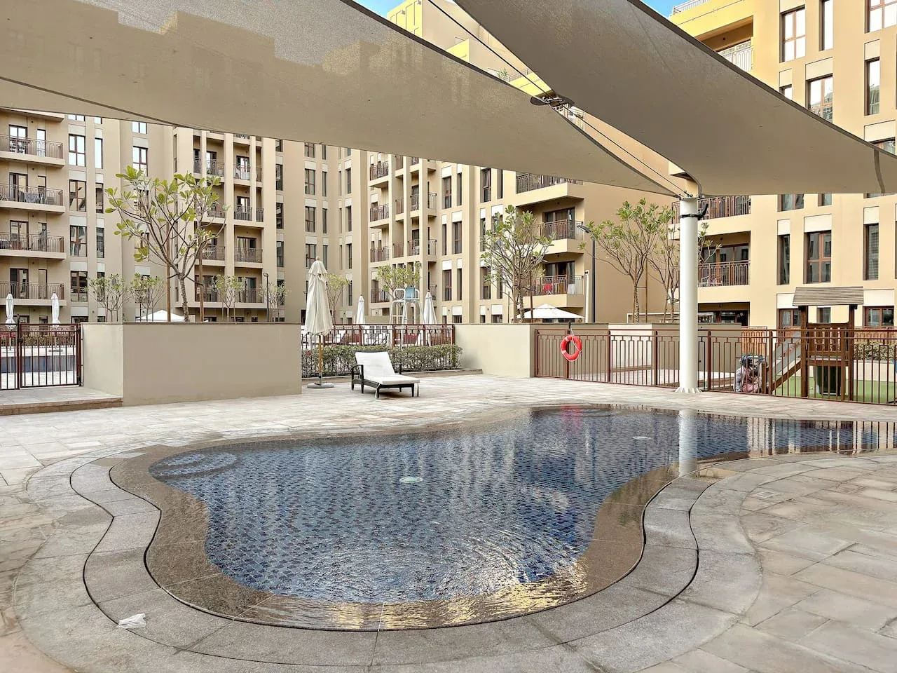 An outdoor swimming pool sits among apartment buildings beneath broad shade canopies.