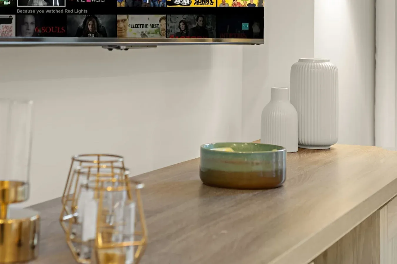 A wooden tabletop displays decorative vases and a small bowl beneath a wall-mounted screen.