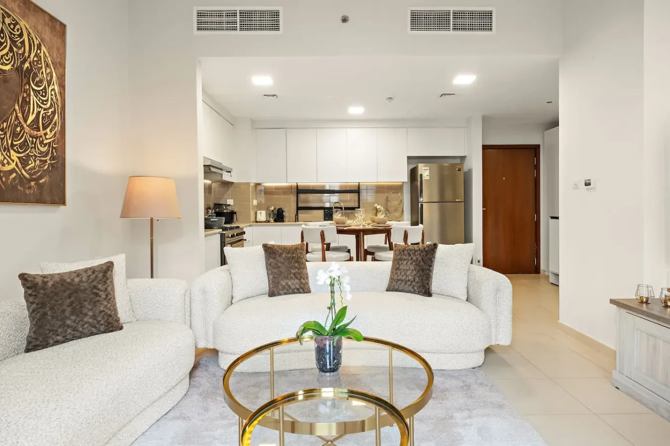 A bright living room with white seating, a glass coffee table, and an open kitchen.
