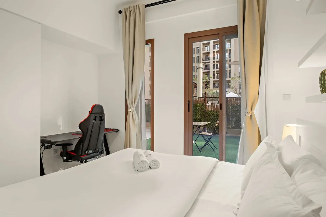 A bright bedroom with white bedding, a desk, and glass doors opening to a balcony.