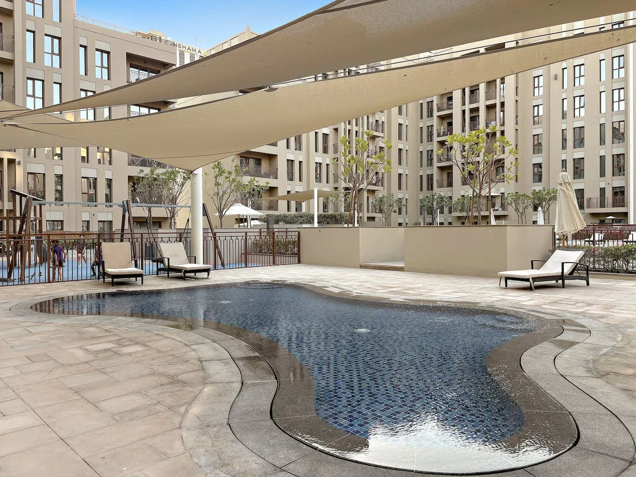 An outdoor swimming pool sits beneath shade sails in a courtyard surrounded by buildings.